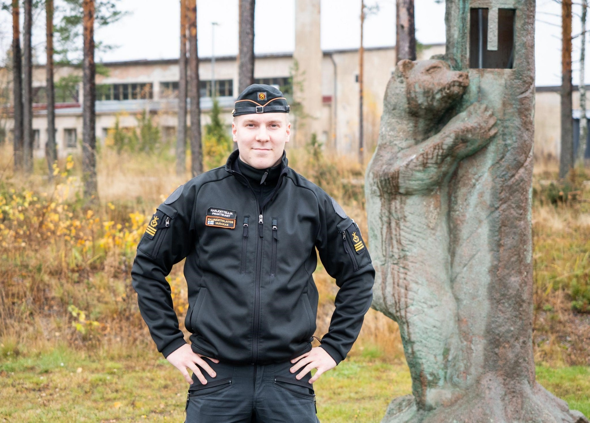 Kadettiopiskelija seisoo metsäisen maiseman edustalla ja katsoo hymyillen kameraan. Taustalla näkyy karhupatsas. 