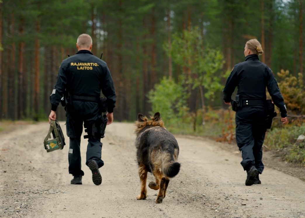 Kaksi rajavartijaa ja rajakoira kävelevät metsätiellä.