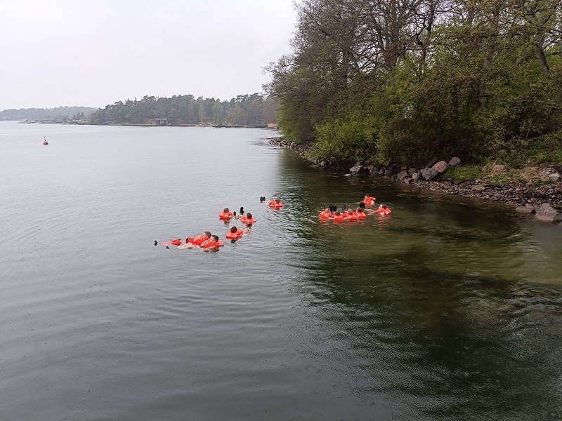 Ryhmä ihmisiä vedessä rannan tuntumassa kellukkeet päällä. Puissa on lehtiä.