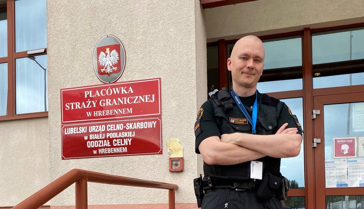 Gränsbevakaren står i sin uniform med armarna i kors framför byggnadens glasdörrar. På väggen vid sidan av namnskyltarna hänger Polands örnvapen.