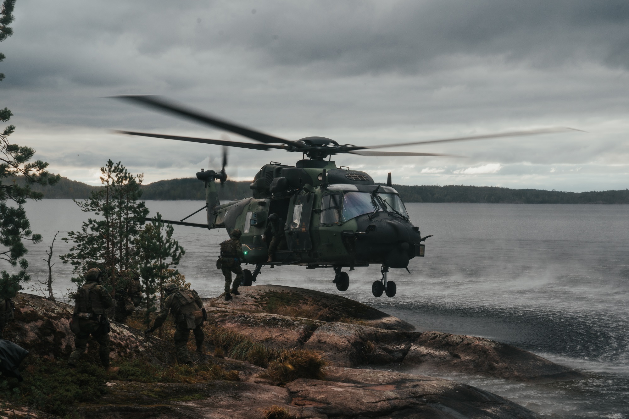 En grön helikopter landar på en klippa. Det finns ett hav i bakgrunden.