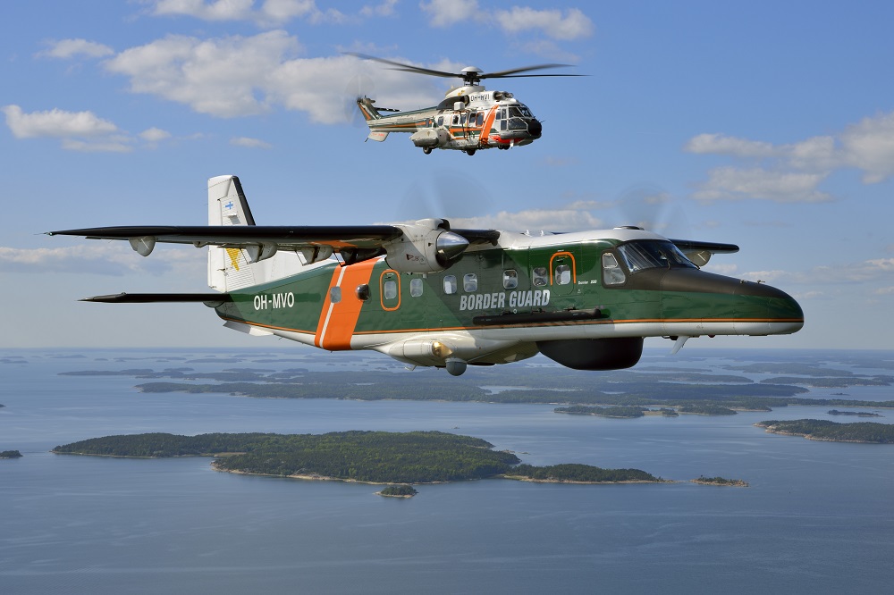 Vihreä lentokone ja sen yllä helikopteri lentää saaristomaiseman yllä kesällä.