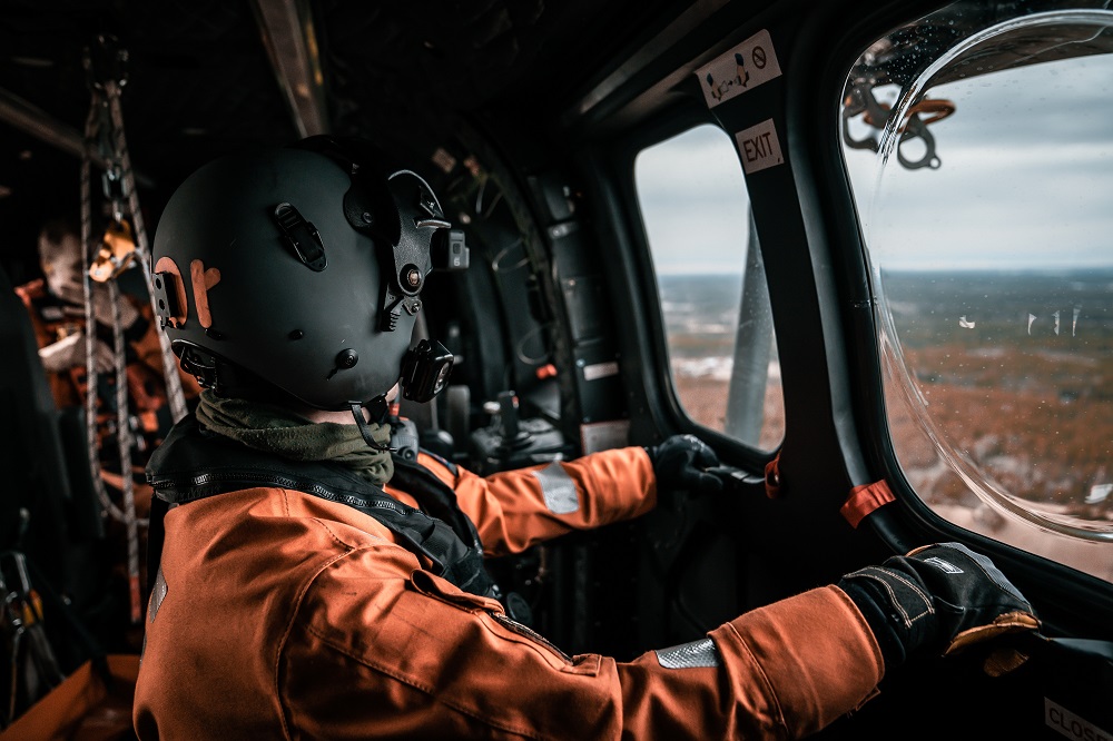 Helikopterin sisällä henkilö oranssissa haalarissa ja mustassa kypärässä katsoo ikkunasta ulos.