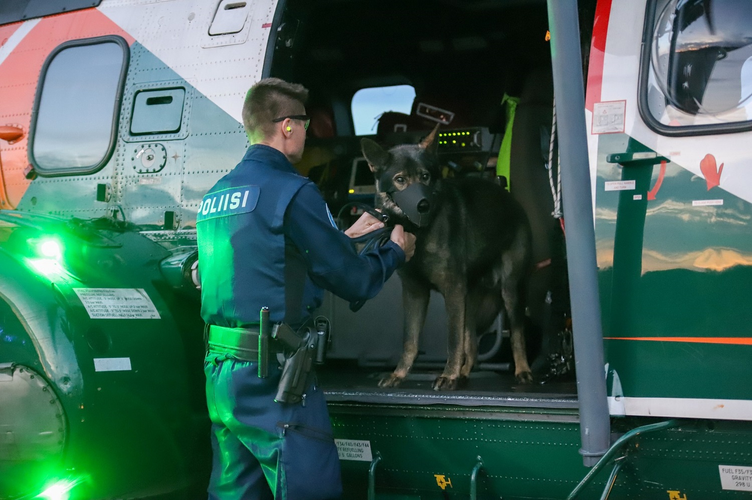 Poliisikoira helikopterin sisällä, poliisi seisoo edessä ja pitää koirasta kiinnni.