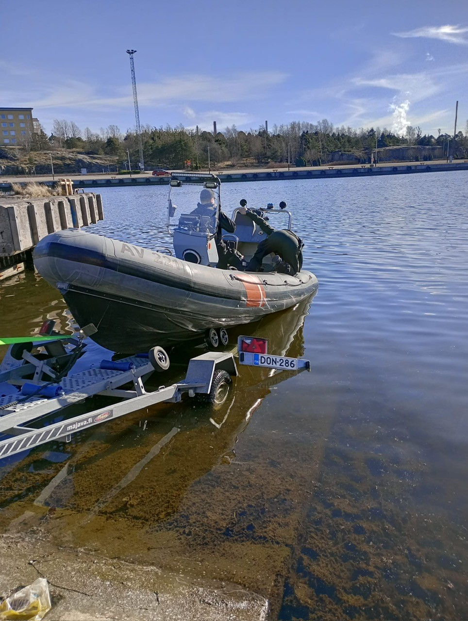 Sjöbevakningssektionens hjälpbåt, en mindre gummibåt, blir lyft upp från vattnet med en trailer. I båten står två tjänstemän med ryggen mot kameran. Vädret är soligt och havet lugnt.
