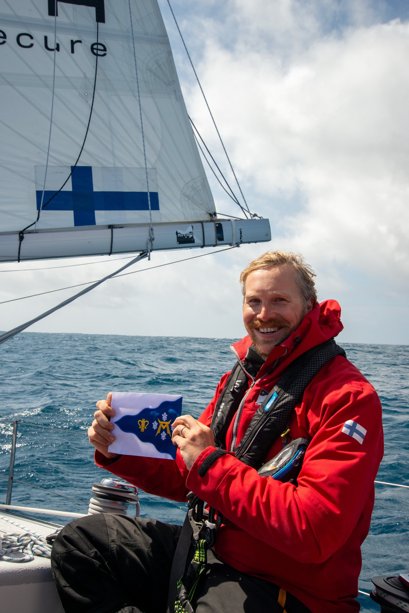 Mikko Hongell istuu purjeveneessä ja pitää kädessään merivartioston lippua. Takana näkyy meri.