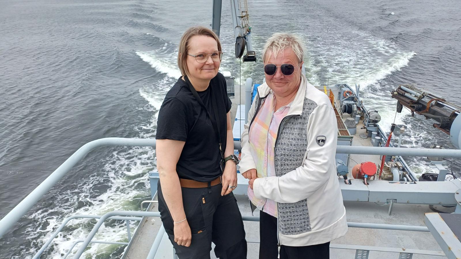 Kuvassa kaksi henkilöä nojaavat laivan ylätasanteen kaiteeseen ja katsovat hymyillen kameraan.