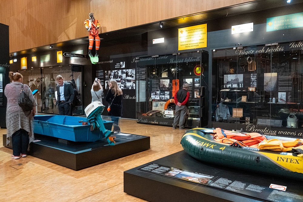 Museon näyttelysali, jossa kaksi pientä venettä, takaseinällä näyttelyvitriinejä ja katosta roikkuu pintapelastaja. Ihmiset katsovat näyttelyesineitä.