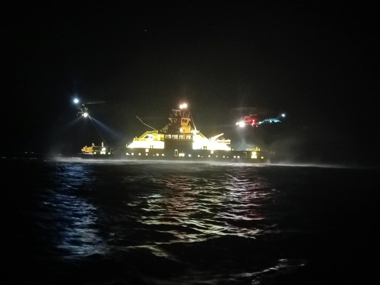 Vartiolaiva pimeällä merellä, yläpuolella kaksi helikopteria vinssaamassa.