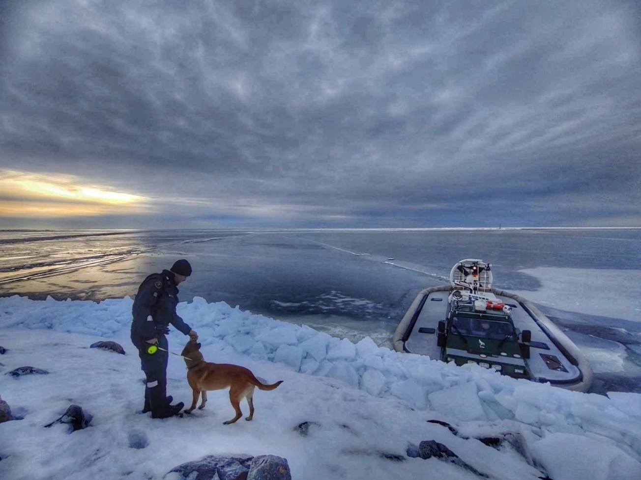 Ett vinterlandskap med sjöbevakare med en hund på isen i förgrunden och en luftkuddefarkost i havet i bakgrunden.