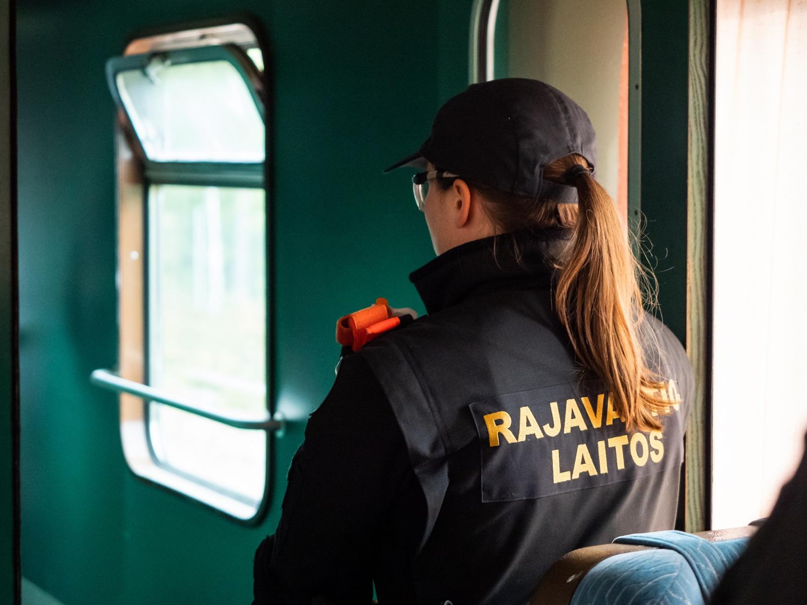 Pitkähiuksinen henkilö kuvattuna takaapäin. Henkilön takissa lukee rajavartiolaitos ja päässä lippalakki. Taustalla näkyy ikkunoita.