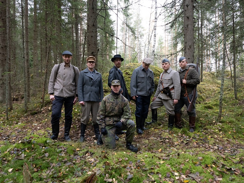 Kuusi henkilöä rinnakkain ryhmäkuvassa, edessä yksi polvillaan. Henkilöt ovat metsässä.