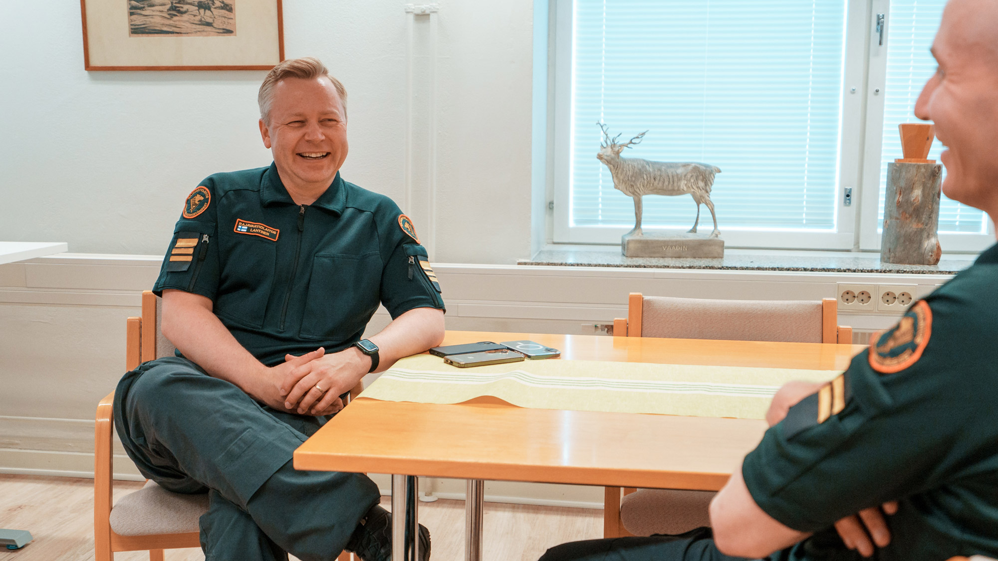 Kaksi Rajavartiolaitoksen univormuihin pukeutunutta työntekijää istuvat kahvipöydässä ja nauravat.