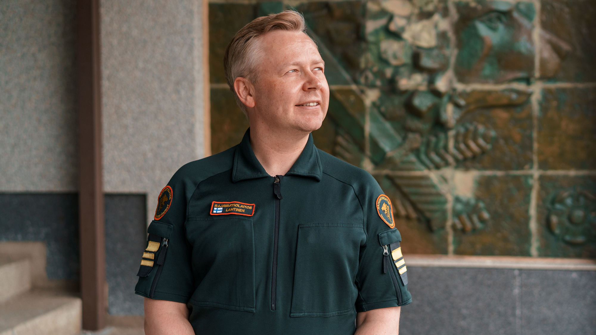 Rajavartiolaitoksen univormuun pukeutunut työntekijä katsoo hymyillen hänestä katsottuna vasemmalle yläviistoon.