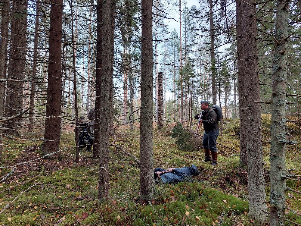 Mies seisoo haulikko kädessä metsässä. Maassa makaa ihminen. Puun takaa näkyy kaksi muuta ihmistä.