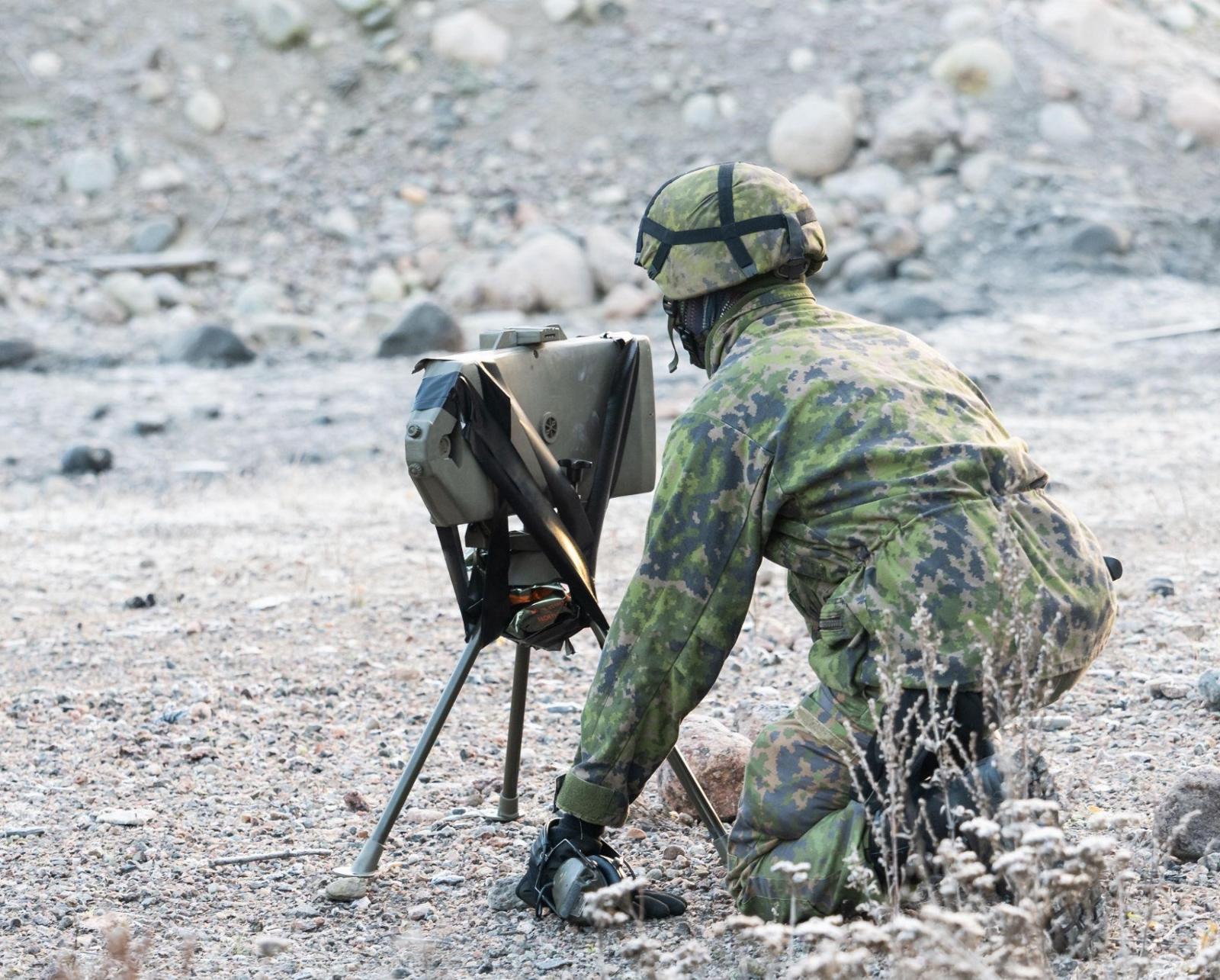 Maastopukuinen henkilö on polvillaan maassa kolmijalkaisen pienen laitteen takana.