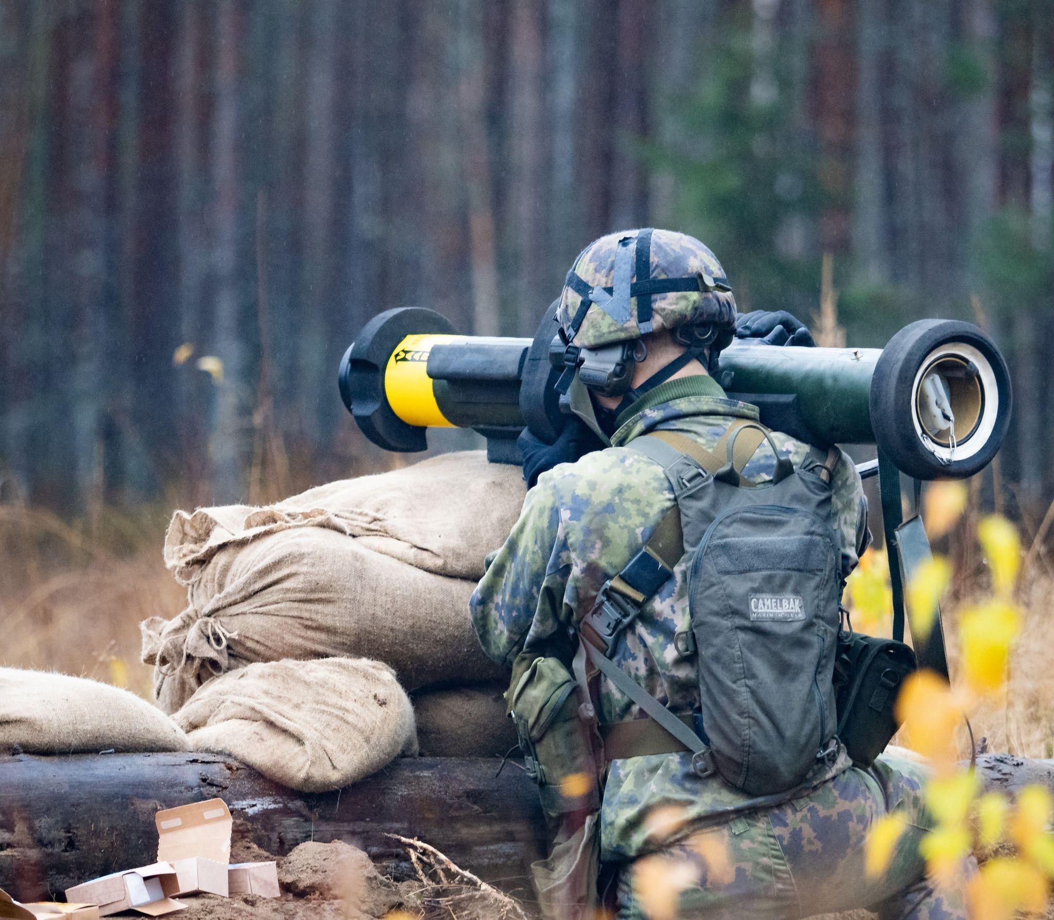 Maastopukuinen henkilö nojaa esteeseen ja pitää olkapäällään sinkoa. Taustalla syksyistä metsää.