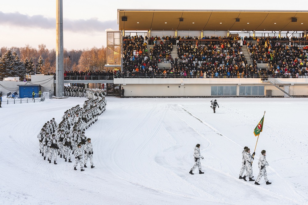 Urheilustadion jossa marssii valkoisiin maastopukuihin pukeutuneita varusmiehiä. Katsomossa istuu ihmisiä.