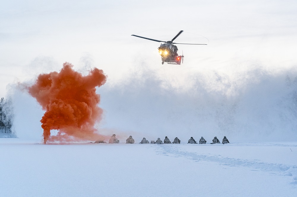 Talvinen maisema, jossa maasta tulee oranssia savua, varusmiehet ovat rivissä matalana ja heidän yläpuolella lentää helikopteri.