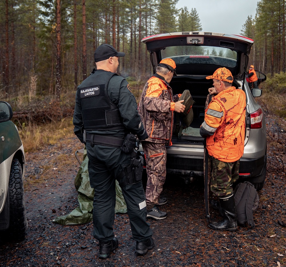 Rajavartija ja kaksi metsästäjää seisovat auton takana. Takaluukku on auki ja metsästäjillä on aseet käsissään. Taustalla näkyy syksyinen metsä.