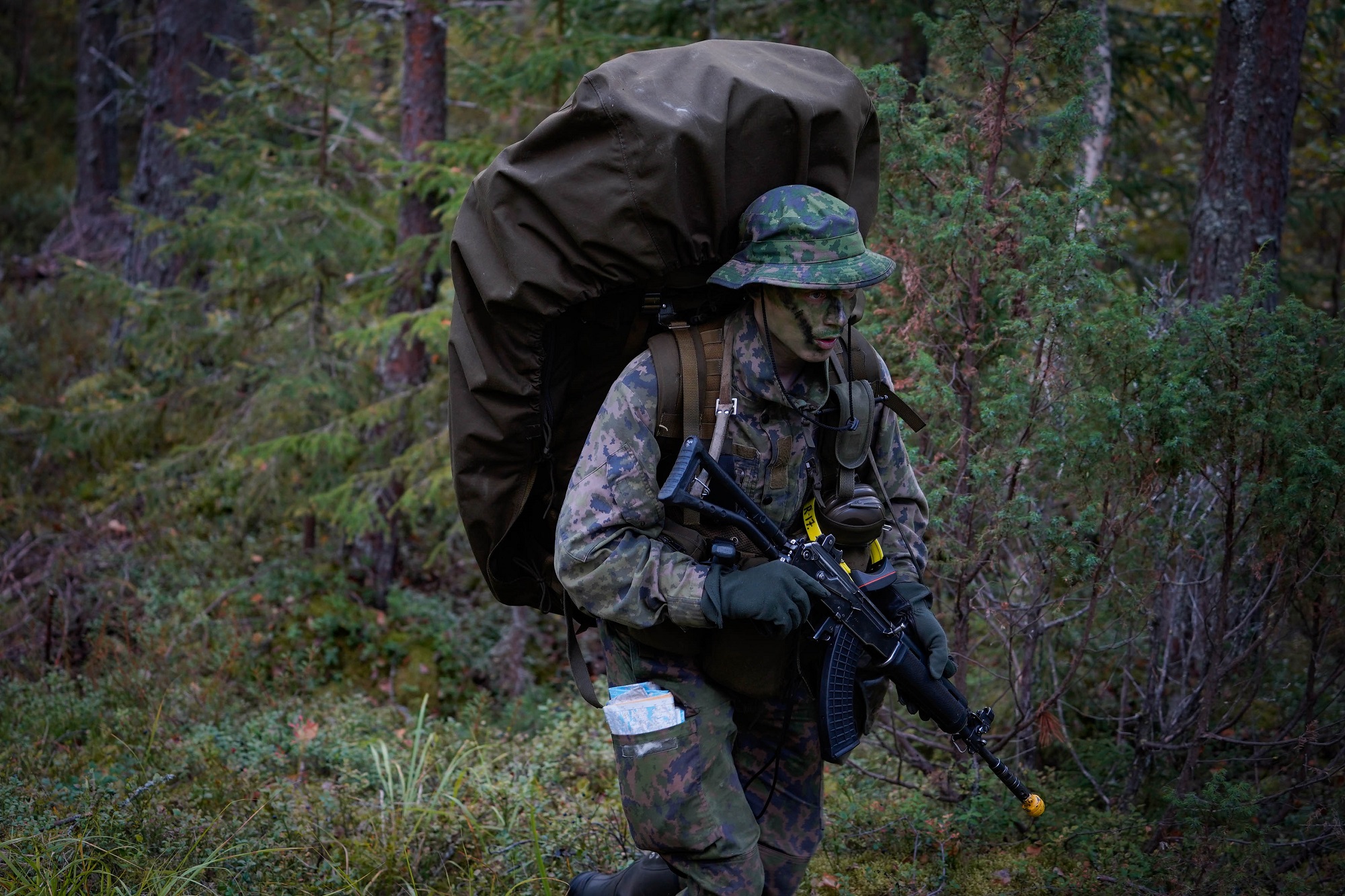 Varusmies metsässä iso reppu selässä ja ase kädessä.