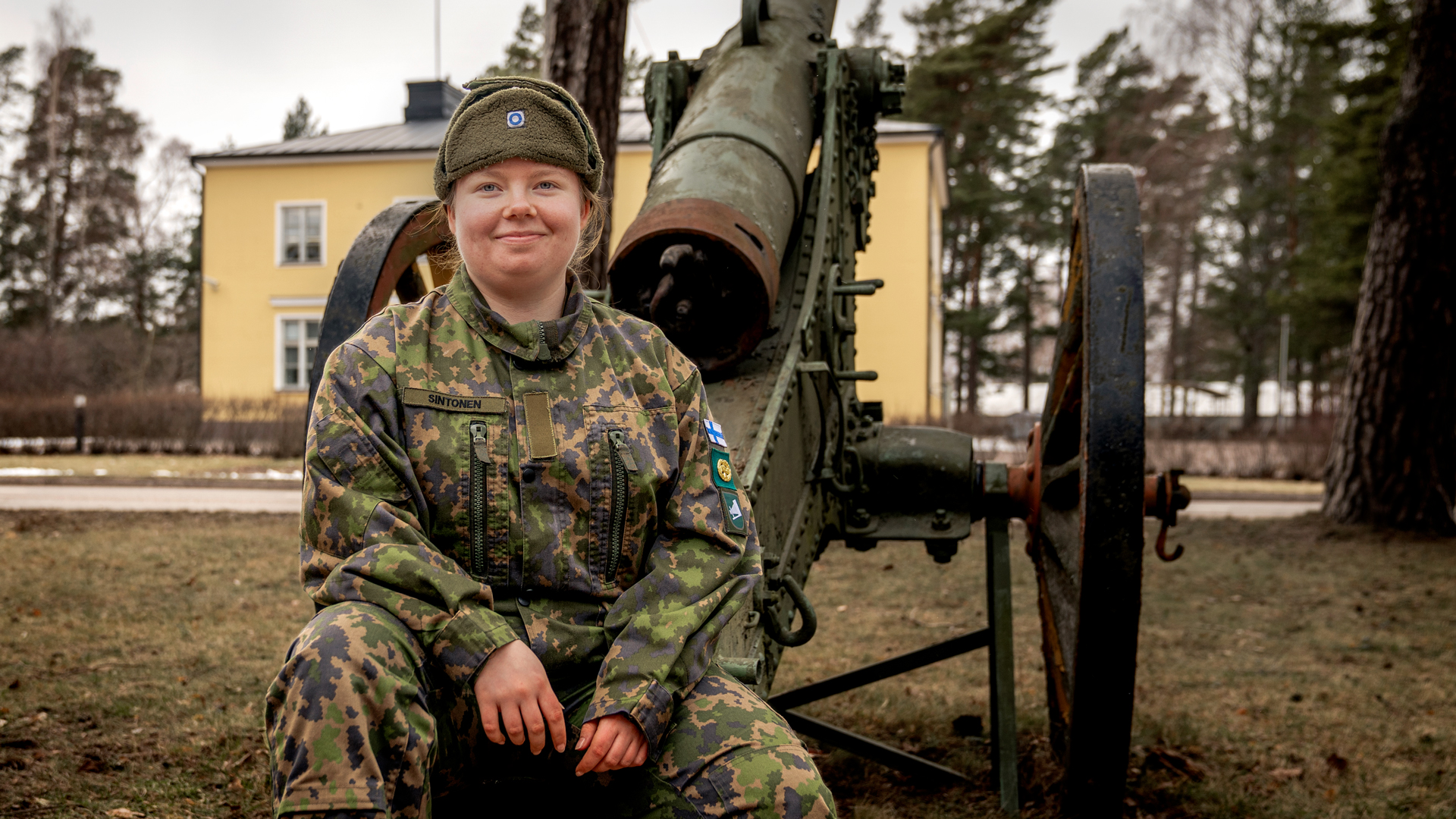 Viestintävarusmies Roosa Sintonen istuu kasarmilla vanhan tykin edessä. Takana keltainen kivirakennus. Kuva otettu aikaisin keväällä.