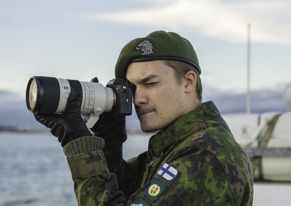 Lähikuva viestintävarusmiehestä keskittyneenä kuvaamaan. Taustalla merellinen maisema.