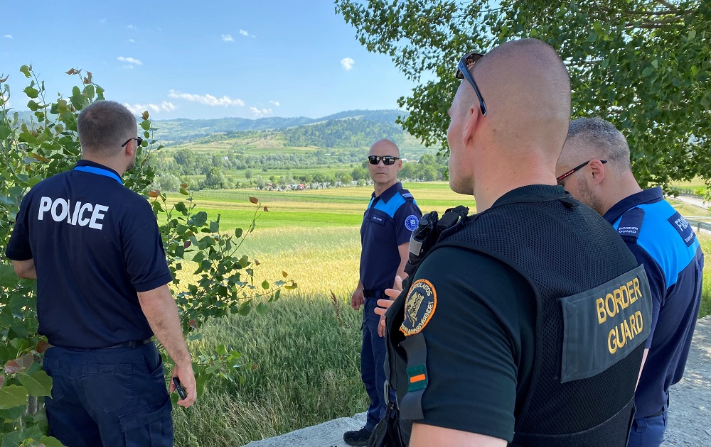 Neljä rajavartijaa keskustelemassa pellon reunalla. Taustalla näkyy vuoristoista maisemaa kesällä.