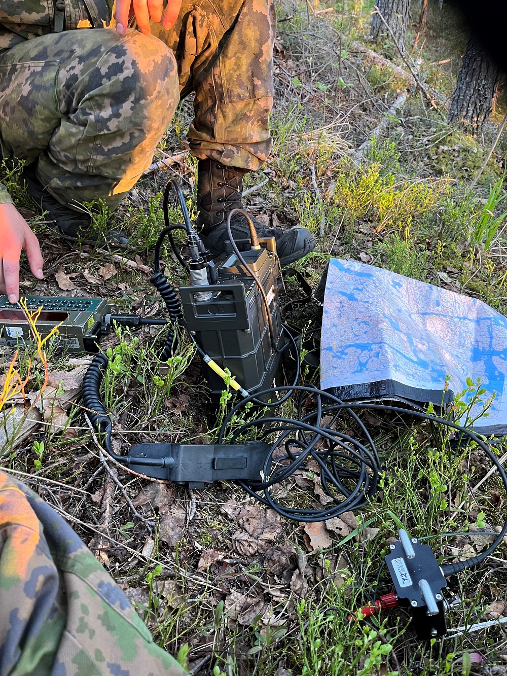 Metsämaasto, jossa maassa erilaisia viestivälineitä ja kartta. Maastopukuisen henkilön jalat näkyvät kuvan reunassa.