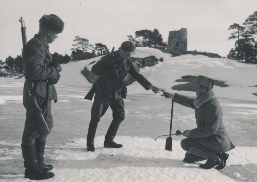 Mustavalkoinen valokuva jossa kaksi rajavartijaa tarkastaa pilkkijän lupaa talvisessa maisemassa.
