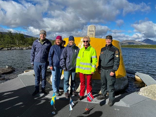 Viisi henkilöä seisoo keltaisen kiven edessä. Taustalla järvi ja tunturimaisema. Etualalla pienet Ruotsin, Suomen ja Norjan liput.