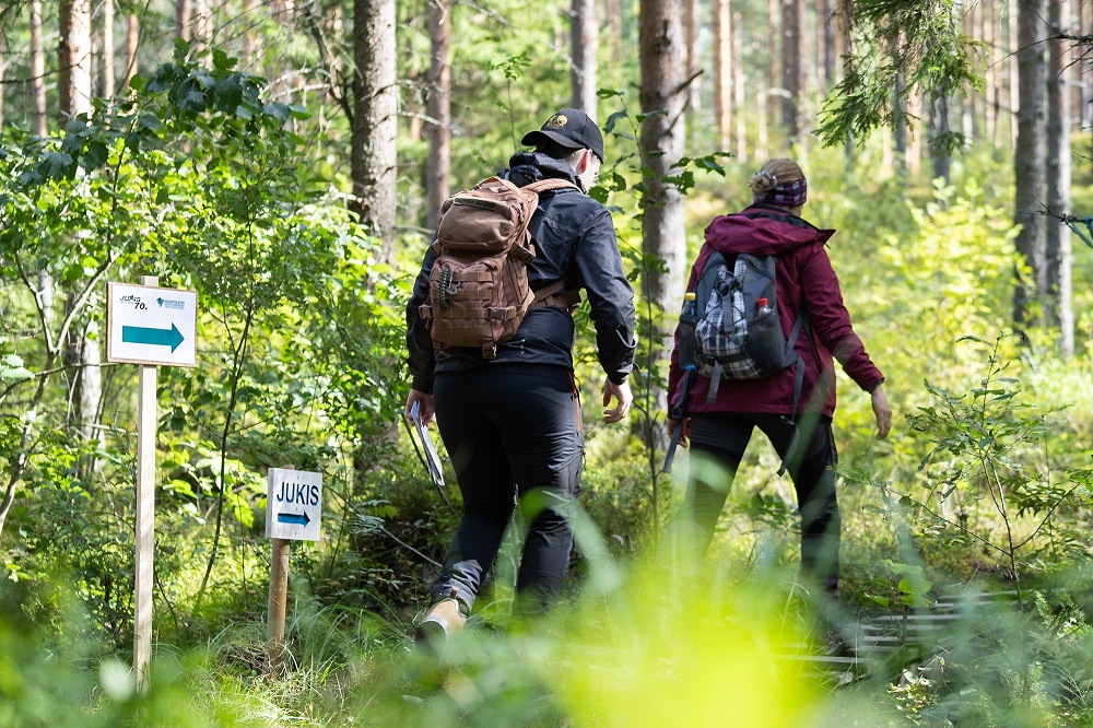 Kolme ihmistä kävelee metsäpolulla peräkkäin. Maasto on vihreää ja kesäistä. Kuvassa kylttejä, joissa lukee Jukis.