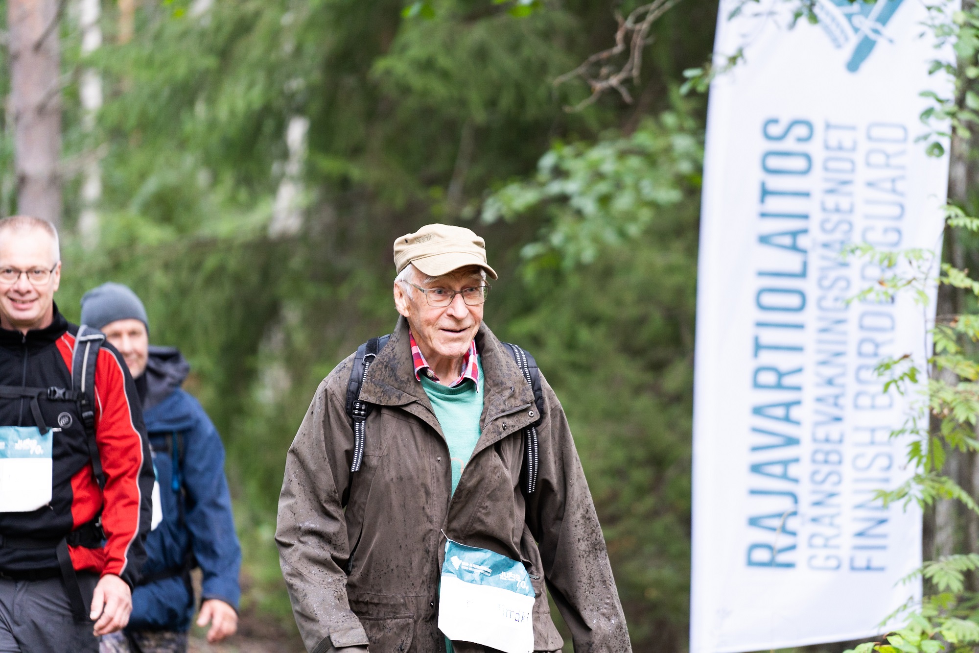 Kolme ihmistä kävelee metsässä. Vieressä Rajavartiolaitoksen valkoinen banneri.