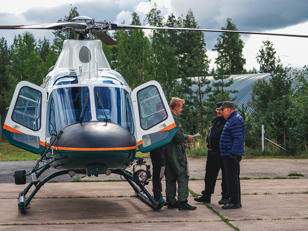Helikopteri laskeutuneena. Sen vieressä seisoo neljä henkilöä jotka keskustelevat. Taustalla puita ja peltinen rakennus.