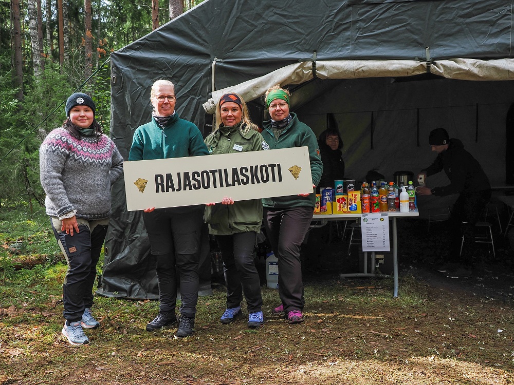 Neljä henkilöä vierekkäin metsässä. He pitävät käsissään kylttiä, jossa lukee rajasotilaskoti. Takana iso teltta, jossa pöytä täynnä elintarvikkeita.