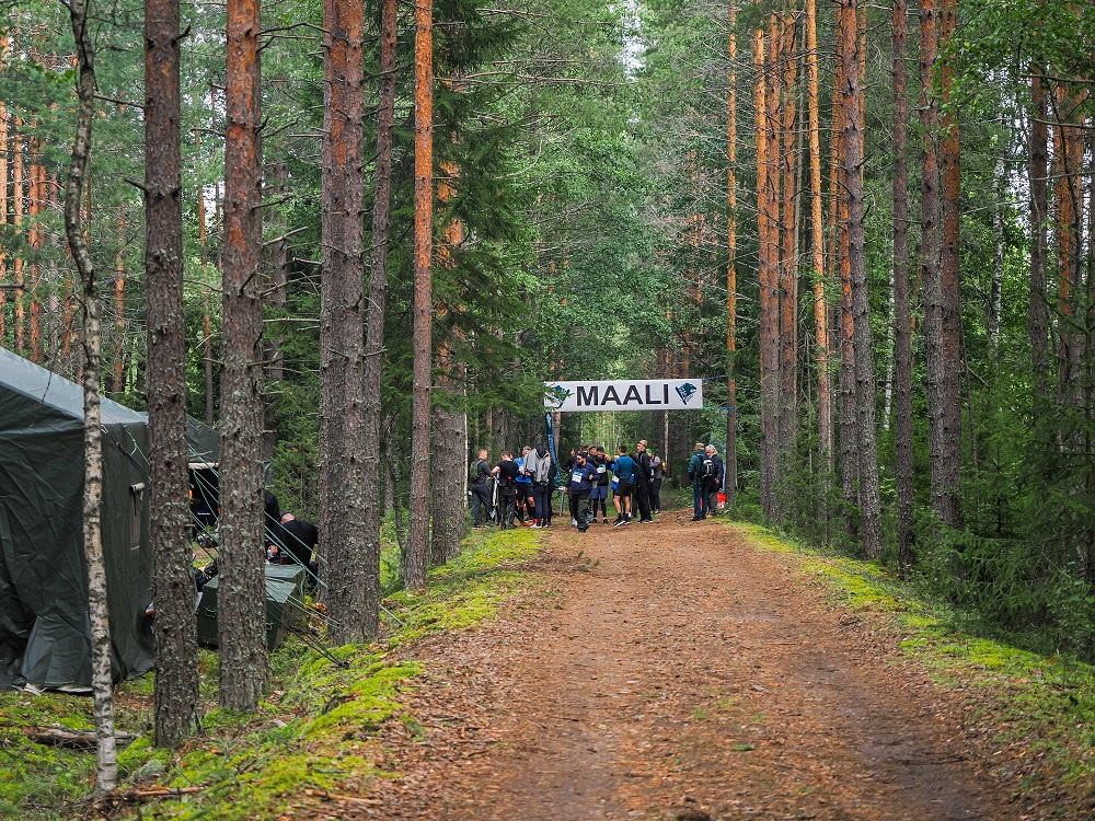 Syksyinen metsä, jossa puiden välissä maalikyltti. Sen alla joukko ihmisiä.