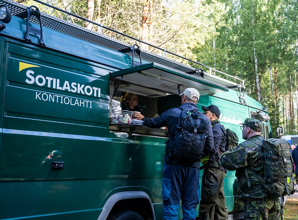 Kome osallistujaa reppuinen asioi sotilaskodin autolla, jossa on kioskimainen luukku.