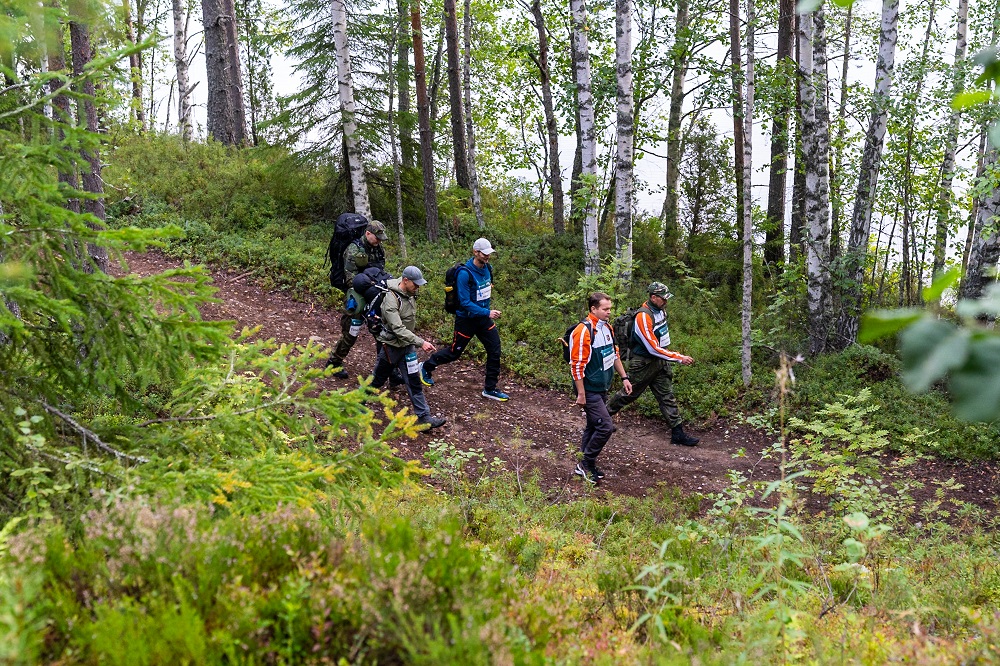 Viisi henkilöä kulkee polulla rinteessä, taustalla puita ja vesistö.