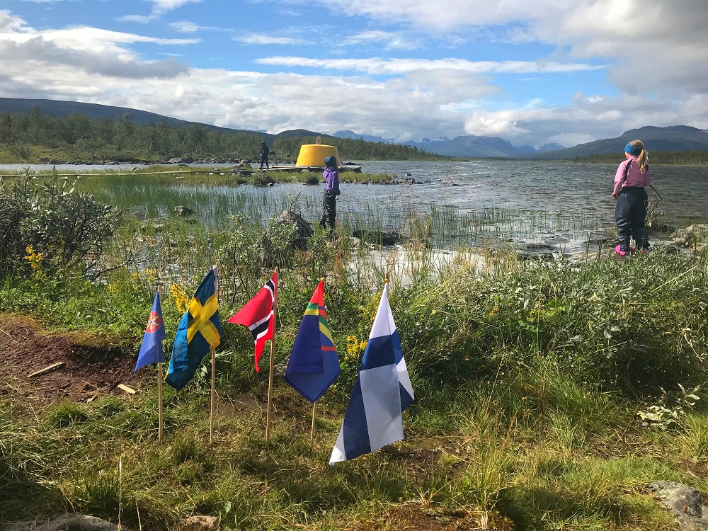 Järven ranta tunturissa. Taustalla keltainen rajapyykkikivi. Kuvassa kolme henkilöä. Etualalla pienet Ruotsin, Norjan, Saamen ja Suomen liput.