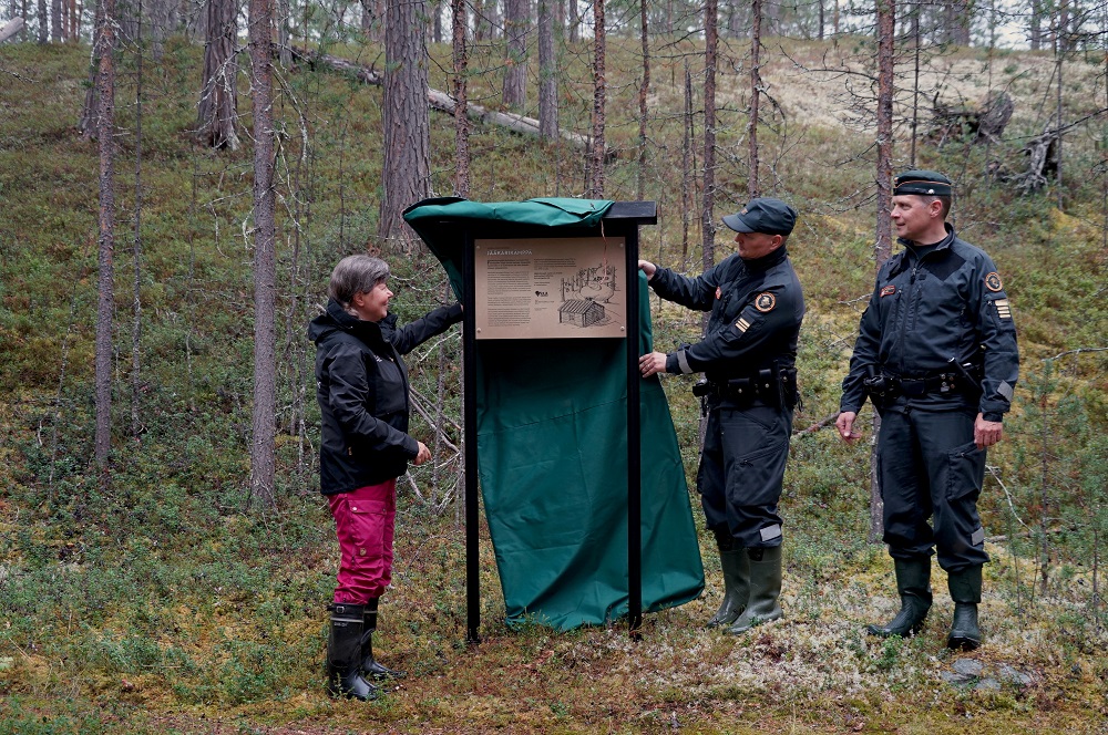 Kolme henkilöä seisoo metsässä ja paljastaa opastekyltin.