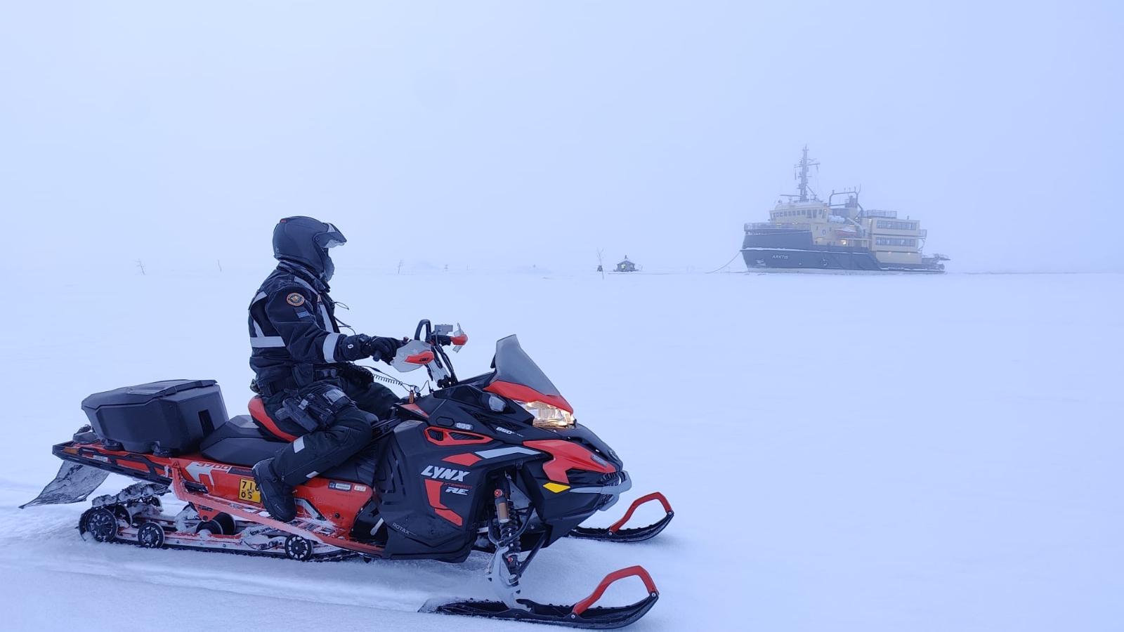 Merivartija katsoo laivan suuntaan moottorikelkan selältä lumisateessa.