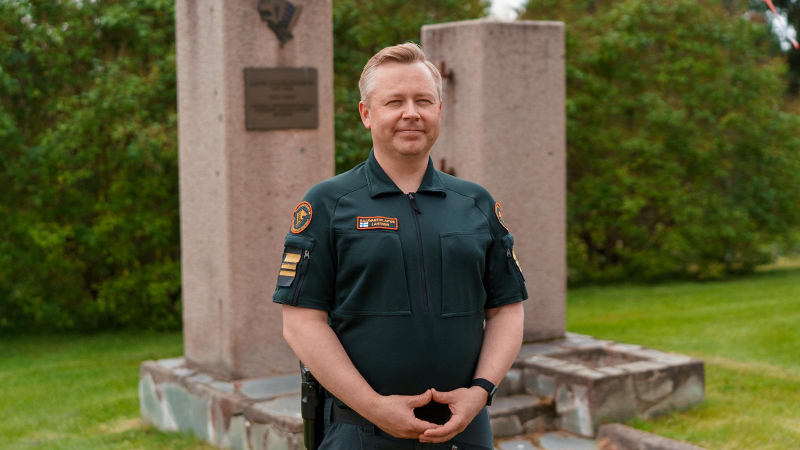 Rajavartiolaitoksen univormuun pukeutunut mies seisoo kuvan keskellä ja katsoo kameraan hymyillen. 