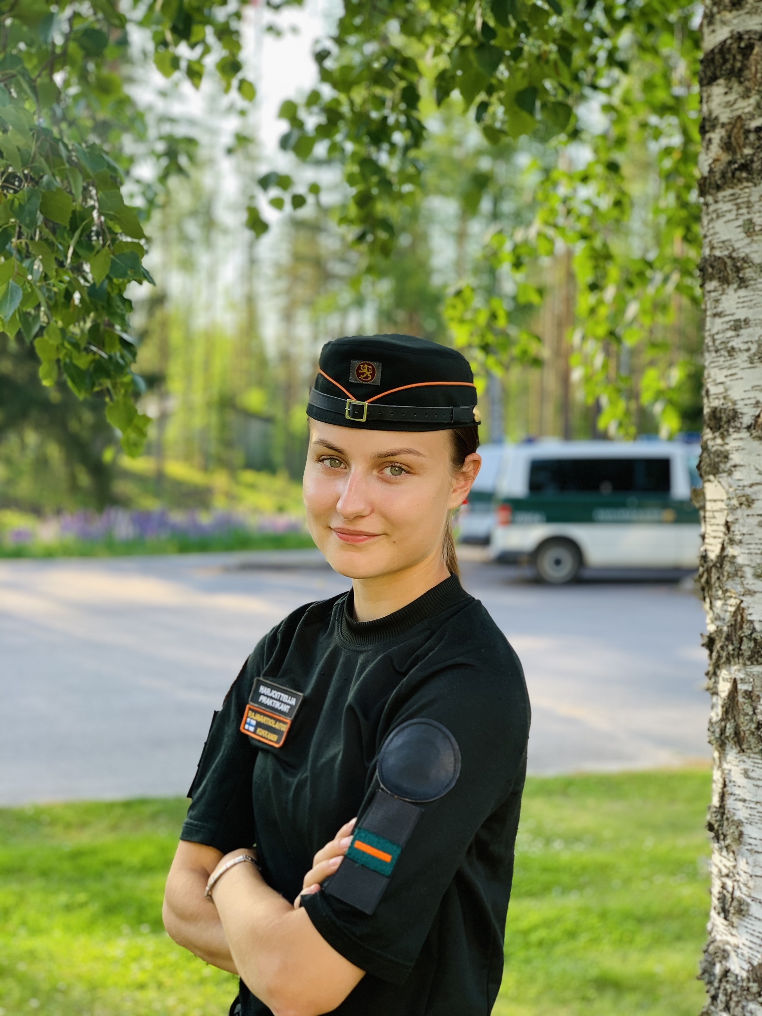 Gränsbevakningsstuderande Suvi Kukkanen i uniformen på sommaren, till vänster en björk, en tjänstebil på bakgrund.